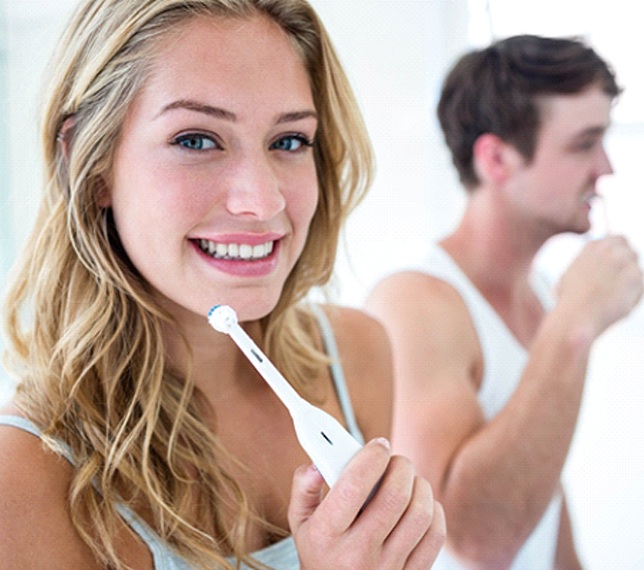 Woman smiling while brushing her teeth with man nearby